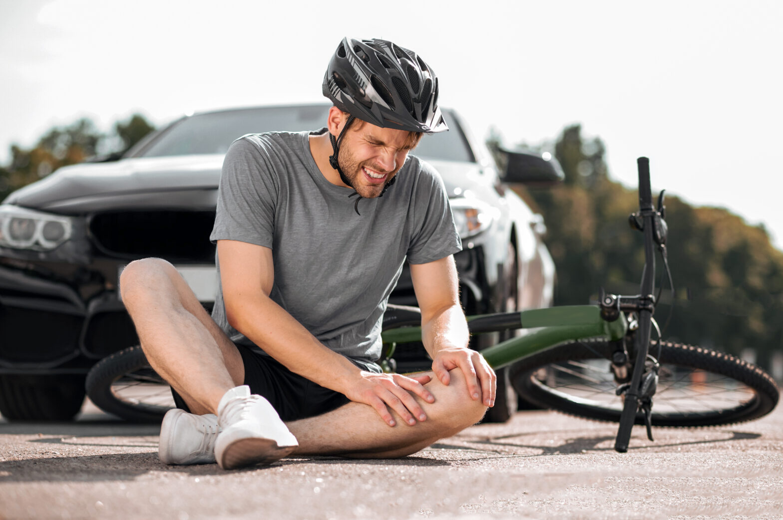 Ein Fahrradfahrer sitzt nach Fahrradunfall mit einem Auto auf der Straße und hält sich das Knie. Im Hintergrund liegt ein grünes Fahrrad und eine schwarzes Auto steht dahinter.